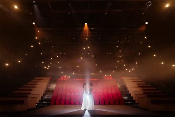 the stage and auditorium with red seats with a couple standing on the stage facing eachother holding hands in white dresses
