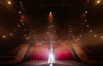 the stage and auditorium with red seats with a couple standing on the stage facing eachother holding hands in white dresses