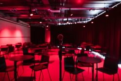A space to hire set up with red lighting and lots of small tables with chairs around each and black curtains along the right hand side