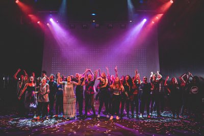 A group standing on stage celebrating. The stage is lit purple and red. The stage floor is covered in confetti.