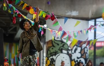 A young person stands in a skatepark adorned with graffiti and bunting, looking up and smiling at a lit lighter in their hand