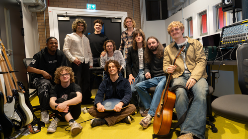 a group of ten men of mixed races and ages sit in a music production studio with a yellow carpet smiling and holding various instruments