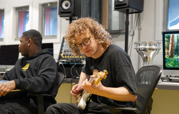 A young white man with mid length curly blond hair and glasses plays a guitar