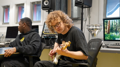 A young white man with mid length curly blond hair and glasses plays a guitar