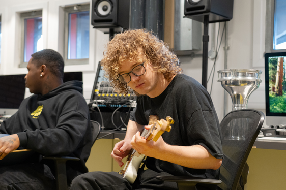 A young white man with mid length curly blond hair and glasses plays a guitar