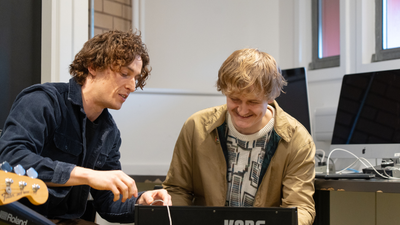 a white man with brown wavy hair shows a young white man with blond hair and a moustache how to play a keyboard
