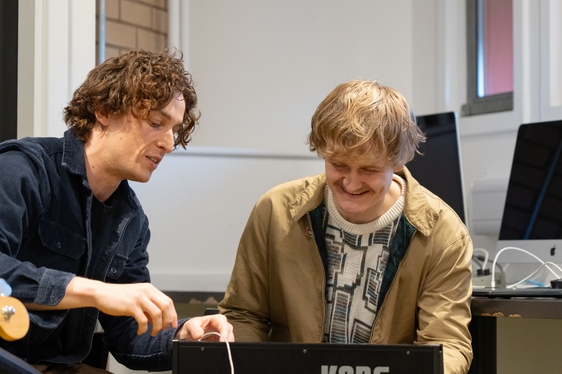a white man with brown wavy hair shows a young white man with blond hair and a moustache how to play a keyboard