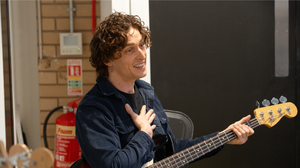 A young white man with medium length brown wavy smiles and hair holds a guitar in one hand and places his other hand on his chest