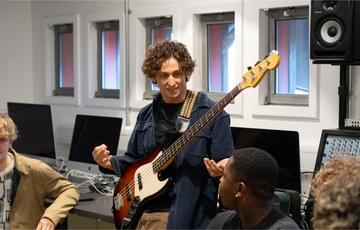 a group of three young men listen to a white man with brown wavy hair man holding a guitar