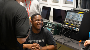 a young black man with short hair and a black tshirt sits in front of music production equipment laughing