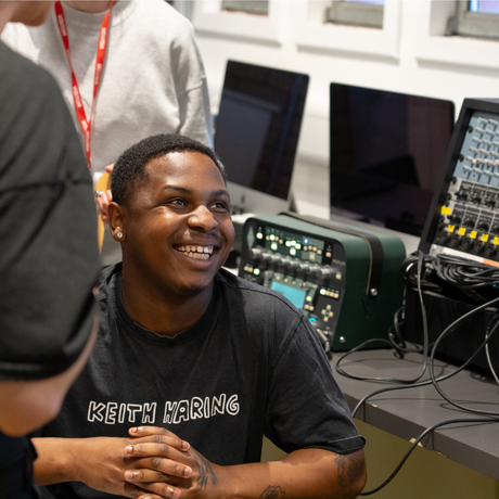 a young black man with short hair and a black tshirt sits in front of music production equipment laughing