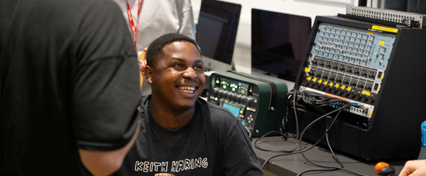 a young black man with short hair and a black tshirt sits in front of music production equipment laughing