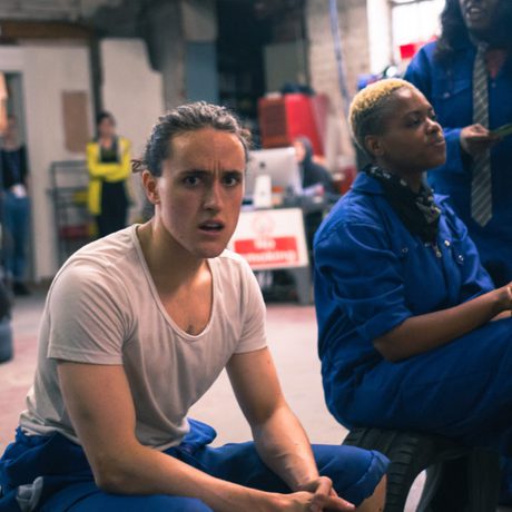 Two people sat on stools in a workshop wearing blue overalls.