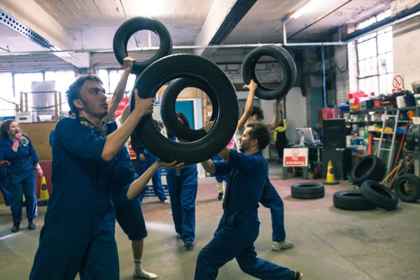 A group of young people wear blue boiler suits performing in a garage