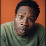 A young black man staring straight into the camera wearing a light green jumper