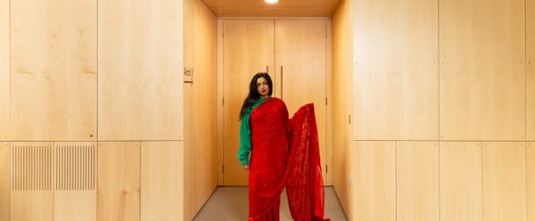 Afreena is stood in front of a modern wooden arched alcove, wearing a bright red sari over a green jumper.