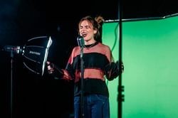 A young woman wears a stripy jumper and sings into a microphone in a studio space