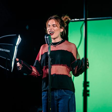 A young woman wears a stripy jumper and sings into a microphone in a studio space