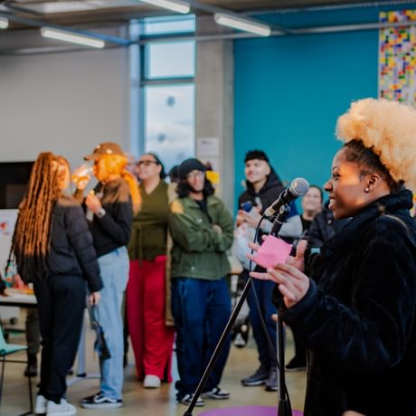A woman with blonde afro hair excitedly addresses a room of people