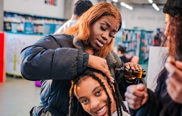 Two young people sat in a library, one working on the other's long braided hair, both smiling