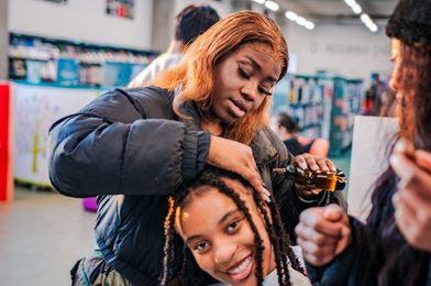 Two young people sat in a library, one working on the other's long braided hair, both smiling