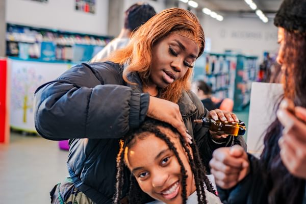 Two young people sat in a library, one working on the other's long braided hair, both smiling