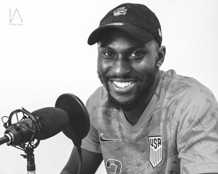 Black and white portrait of Paul Olubayo. He is wearing a cap and smiling with a recording microphone next to him.