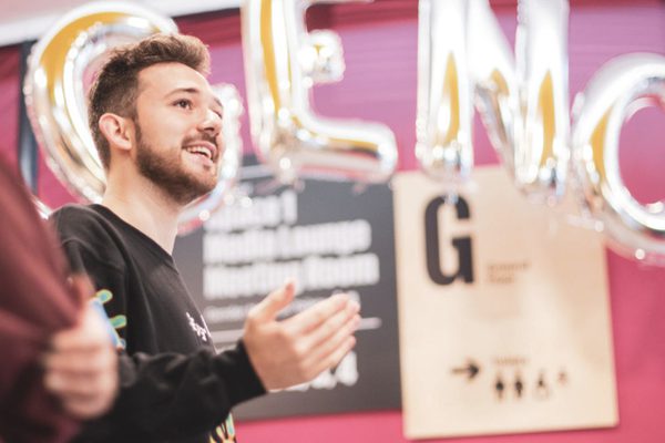 A young man stands under balloons reading AGENCY whilst addressing a room of people