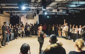 A man stands in the centre of a room speaking passionately to young people