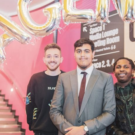 Three young people smile at the camera under balloons that read AGENCY