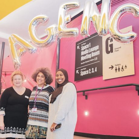 Three people stand on stairs proudly below balloons reading AGENCY