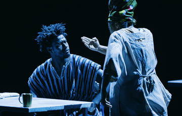 Two performers on stage. They are having a conversation and one of the performers has their back facing the audience. They are lit blue by stage lights.