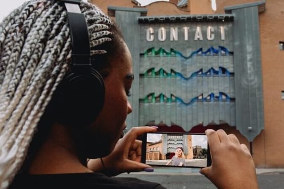 A young person stands in front of Contact, a castle-like theatre building, looking into a phone screen showing a clown outside of the venue